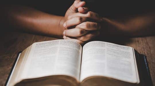 Mãos em formato de prece, em frente a uma bíblia aberta.